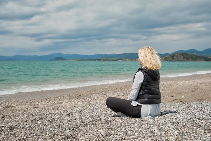 Blonde woman sitting quietly on a pebble beach, reflecting – ADHD coaching for women