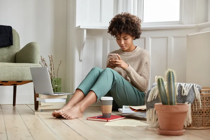 Woman sitting on floor with phone and laptop in calm home setting representing ADHD planning struggles and organisation challenges