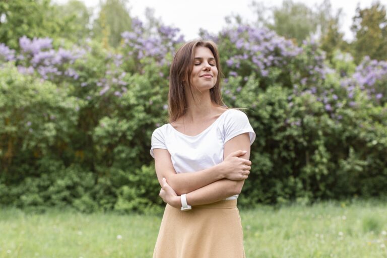 Woman standing peacefully outdoors with arms gently crossed in a self-hug, representing self-compassion and calm.