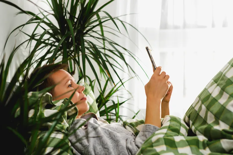 Woman lying in bed scrolling on her phone surrounded by plants, representing ADHD procrastination, task paralysis, overwhelm, and waiting until the last minute to start tasks.