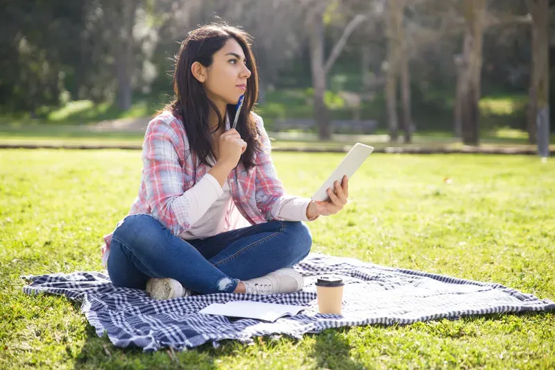 Woman sitting on a blanket in the park with a notebook, reflecting on being only motivated by deadlines with ADHD