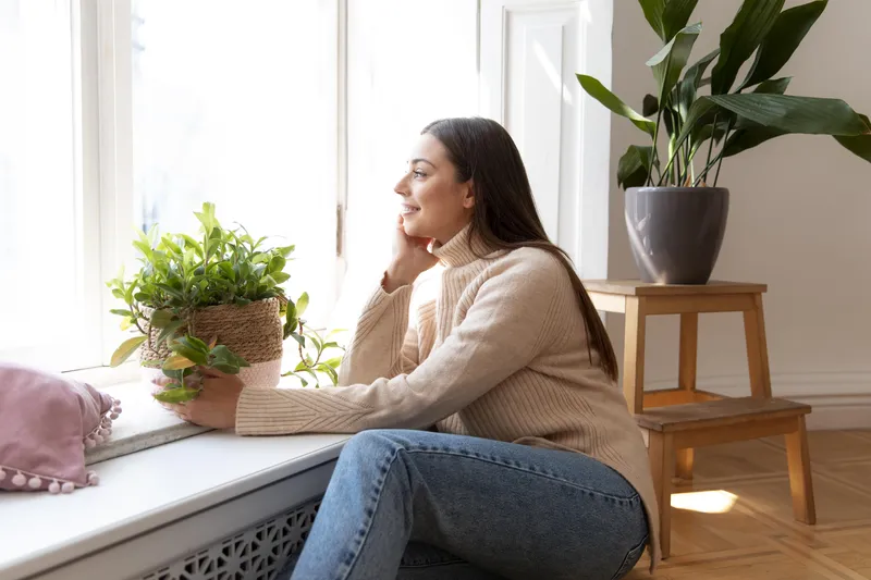 Woman sitting beside window holding plant in calm home setting representing supportive ADHD coaching for overwhelmed adults