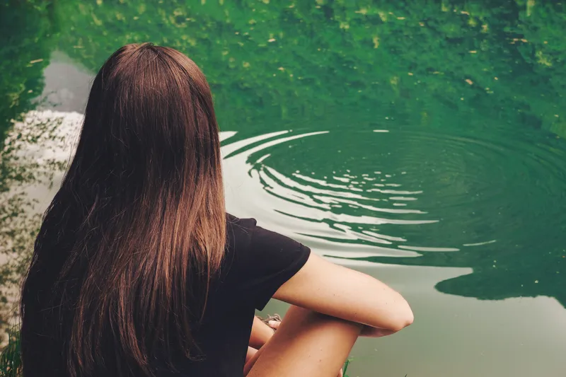 Woman sitting beside calm green water reflecting quietly during ADHD therapy and emotional support work
