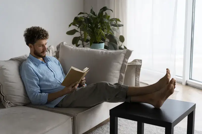 A man sitting on a sofa trying to read a book but lost in thoughts of why getting stuff done with ADHD can be so hard.
